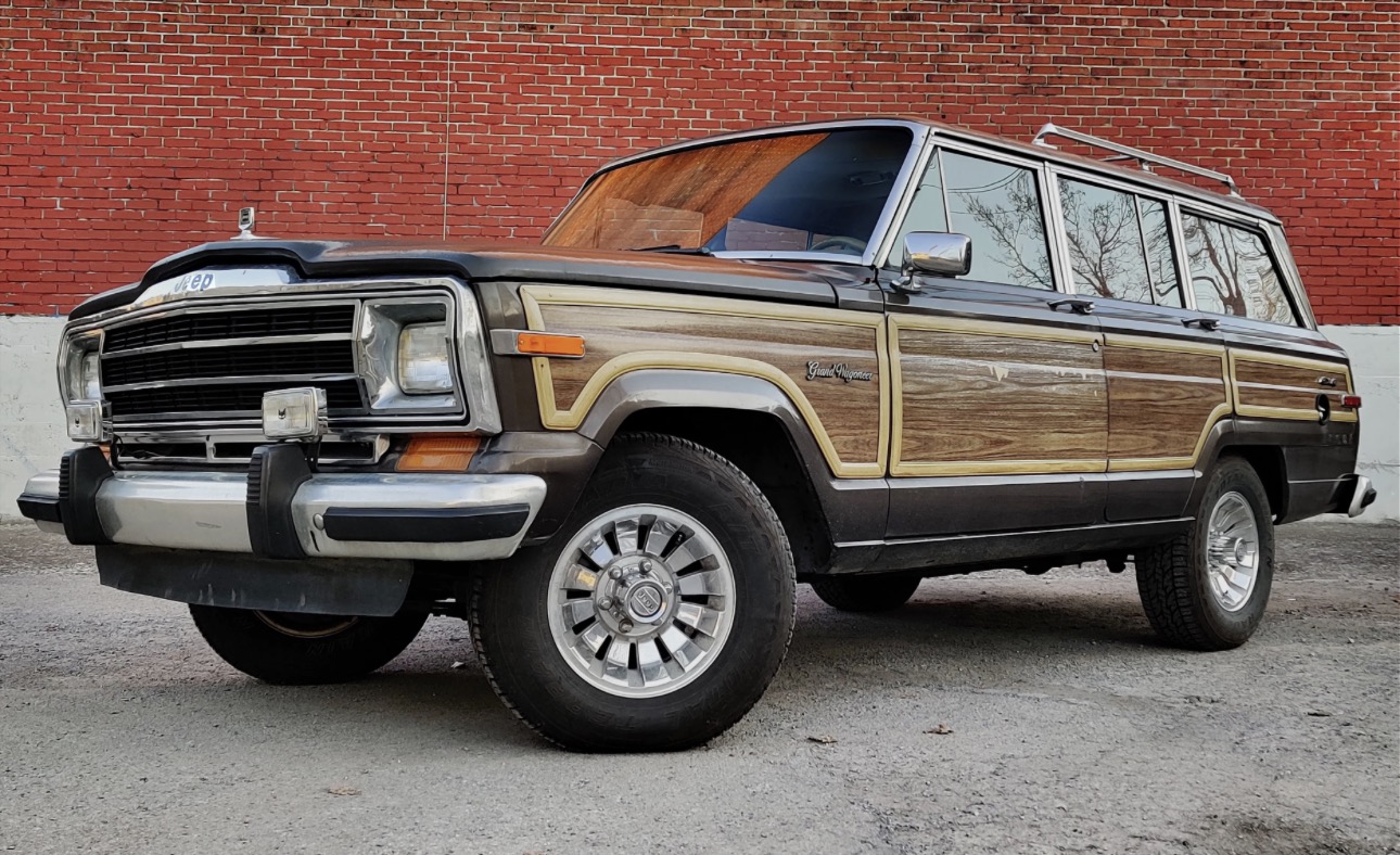 Classic Jeep Grand Wagoneer with woodgrain side panels parked in front of a red brick wall, illustrating an iconic platform for engine swap and restoration options.