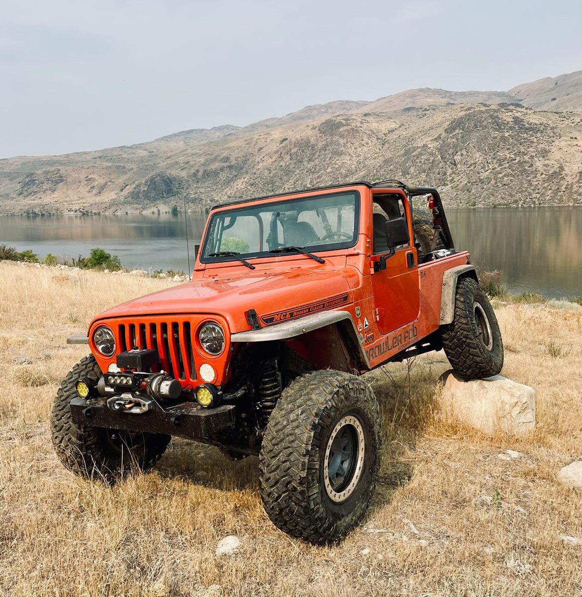 Lifted orange Jeep Wrangler flexing on a rock beside a lake, showcasing an off-road build that highlights different Wrangler engine options.