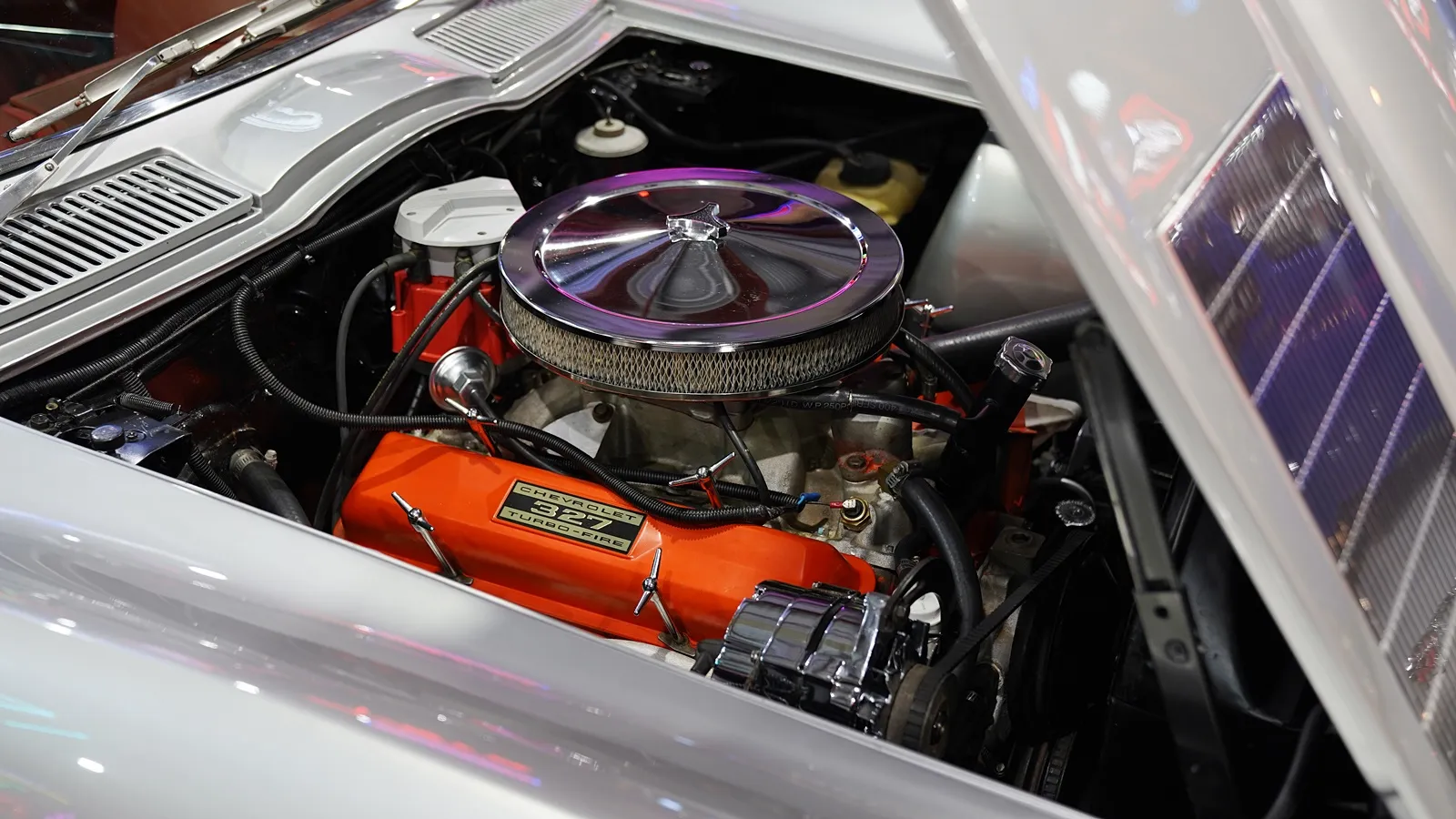 Classic Chevrolet 327 small‑block V8 with orange valve covers and chrome air cleaner under the hood of a vintage Corvette, showcasing a popular builder engine.